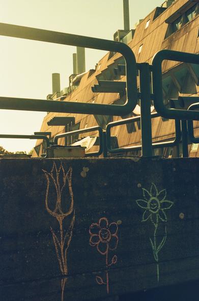 Three chalk flowers are drawn on a concrete wall. One orange tulip, one red and one green flower with a smiling face.

In the background is a brutalistic building, the Mäusebunker.