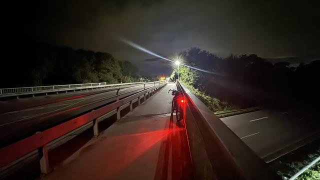 Ein Fahrrad ist nachts auf einer dunken beleuchteten Brücke geparkt, mit roten Lichtern auf dem Fahrrad und der Fahrbahn darunter. Die Umgebung ist dunkel, mit beleuchteten Abschnitten in der Ferne.