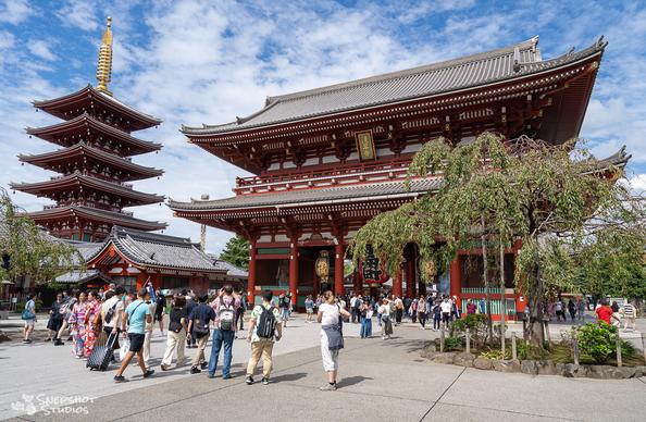 Daytime photo of a red pagoda on the left and a temple gate on the right. Groups of visitors walk around in the open spaces.