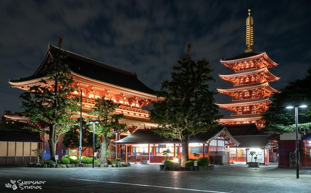 Nighttime photo of a red temple gate on the left and a pagoda on the right. Both illuminated by artificial lights. The space is deserted and no people are visible