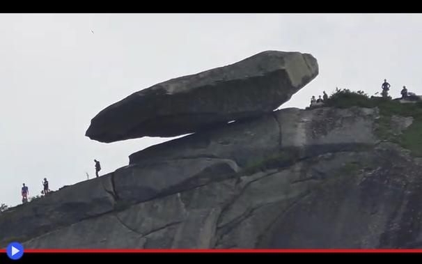 Foto presa da lontano e in controluce di un grosso macigno oblungo in apparente equilibrio precario su un pendio scosceso. Alcune figure umane che vi camminano attorno sembrano in pericolo di venire schiacciate a momenti.