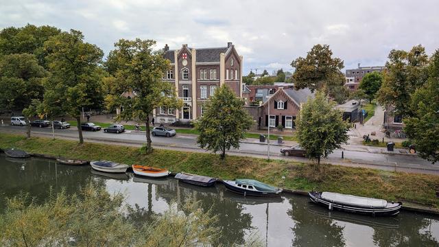 View from my hotel window over a straight section of a tree-lined canal with small boats moored along the side, most of them covered over with tarpaulins. There's a street running alongside the canal, with parked cars, and a cyclist travelling down the cycle lane that runes alongside that. Some beautiful houses beyond that, in a style I can only (poorly) describe as "Dutch" (sorry!). The sky is grey and cloudy.