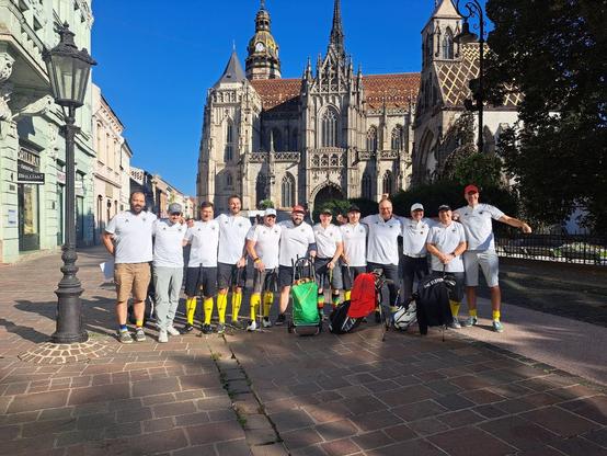 Die deutsche Crossgolf Nationalmannschaft steht in Kosice in der Straße vor der Kirche zur Präsentation.
Alle haben weiße Sporttrickots an und meist kurze Hosen. Viele haben dazu noch gelbe Kniestrümpfe an