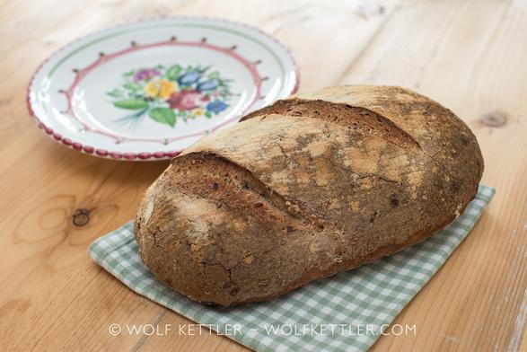On a wooden table top a rustic loaf of bread sits on a green and white checked, folded placemat. In the background a small decorated Austrian plate.