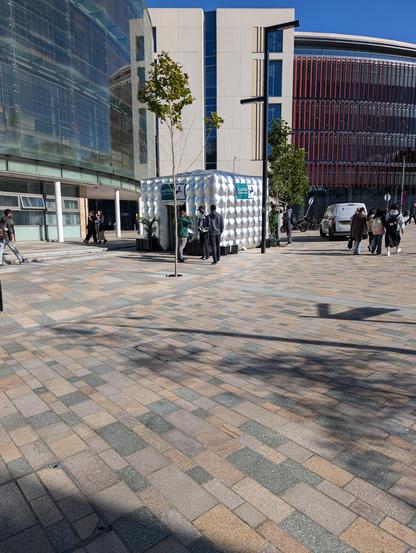 An inflatable cube made of silver material stands on a plaza of pink and grey granite setts. The cube has green, white and black signs on the sides advertising Lloyds Banking Group. Several people in green branded tops are talking to University students. In the background are the curved glass walls and polished concrete walls of modern university buildings.