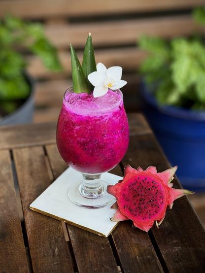 bright vivid magenta cocktail with pineapple frond and white orchid garnish sitting on a white coaster next to the top slice of a dragon fruit with the seeds exposed with chili plants in the background