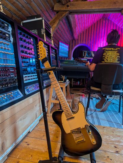 Photo du Studio de Alias avec en premier plan une guitare électrique, quelques rack 19 pouces bien fournis et une console de mixage en arrière plan.