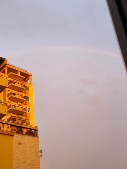 Arcobaleno che spunta da un palazzo, mentre il cielo è coperto e piogiggina