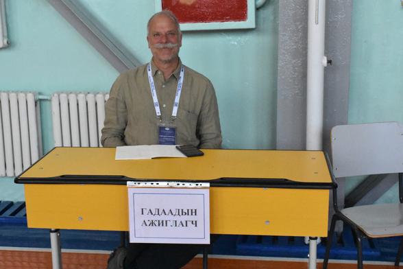 Photo of me during election observation this past June. I'm sitting at a school desk that has a sign on it saying "foreign observer" in Mongolian on it. A set of notes and my cell phone in front of me on the yellowish desk. I'm wearing my observer ID on a lanyard around my neck and smiling serenely! The wall behind me is that ubiquitous (in Mongolia) mint colour.