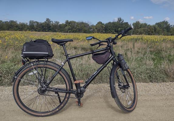 A black bicycle posed on the side of a gravel road.  A field of grasses with yellow flower tops is visible across the photo behind the bike and road.  At the far side of the field a line of trees stretches across the view.  A vivid blue sky is above the trees with a couple small white clouds just in view.