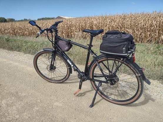 A black bicycle posed on the side of a gravel road.  Past the bicycle is a field of tall corn plants.  All the corn stalks are brown and dry.  Just above the tops of the corn, a small corner of a farm outbuilding can be seen in the far distance. The roof of the building is a light tan color that matches the gravel road.  A light blue sky is visibile above the entire picture.