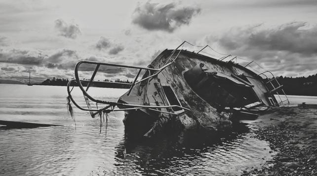 A badly damaged liveaboard boat, beached. The top has been stripped down , there's a giant gaping hole into lower cabin. It's wrecked & tipped on its side, partly buried in sand at high tide line.
Black and white photo.