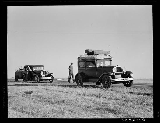 The image is a black and white photograph depicting two vintage Model T-style automobiles parked on an open, grassy field. One car has its roof removed to reveal passengers seated inside. Both vehicles are adorned with luggage atop their roofs, suggesting travel or transportation of goods.

In the background stands a person in work attire: overalls and a hat, seemingly observing or overseeing something related to these cars. The setting appears to be rural, possibly during an early 20th-century timeframe due to the design of the automobiles and clothing style.

The image carries historical context as it depicts scenes from "Between Tulare and Fresno," relating two families originating from Independence, Kansas who are engaged in cotton-chopping work near Fresno that morning. The photograph is attributed to Dorothea Lange, an American photographer known for her documentation of various social issues including the Great Depression's impact on rural communities.

This image captures a moment in time showcasing not only transportation but also labor and migration during early 20th-century America, highlighting historical aspects such as industrialization, travel habits, and economic activities.