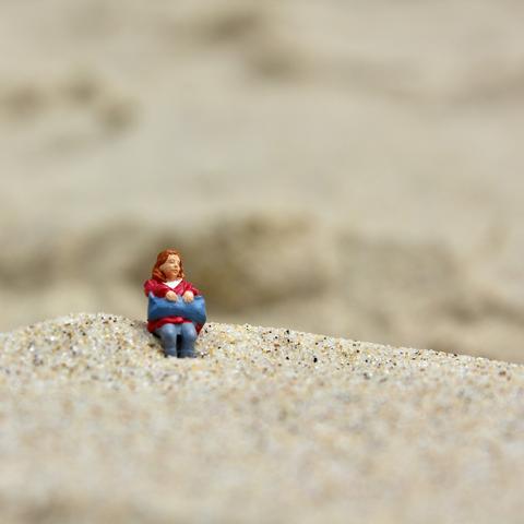 das quadratische bild zeigt aus der froschperspektive eine modelleisenbahnfigur, eine sitzende frau mit einer blauen tasche auf den beinen, im sand sitzend.
die nahaufnahme hat einen engen fokusbereich. die figur und die mini-düne, auf der sie sitzt, sind scharf im fokus und leicht von unten nach oben fotografiert. der bereich davor und der ganze hintergrund sind unscharf als sand womöglich an einem strand wahrnehmbar.
die figur macht einen wartenden, gelangweilte eindruck.