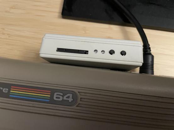 La imagen muestra un emulador de unidades de disco SD2IEC. Es una caja de plástico conectada al commodore 64, ocupando el puerto serie y el puerto del casette, con dos pilotos LED y dos botones negros a la derecha de una ranura para tarjetas SD.