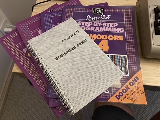 En la imagen está el manual oficial del Commodore 64 abierto por el capítulo 3, “BEGINNING BASIC”. Bajo este manual están los 4 libros de la serie “Step By Step Programming Commodore 64” de Phil Cornes.