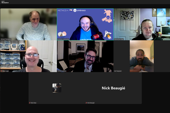 This image shows a Zoom virtual meeting with eight participants, six of whom have their cameras turned on, while two have their video off.

The person on the top left is named Richard Thompson, who appears to be sitting in front of a blurred background while wearing a grey sweater.
The center participant, with the name "Owain Jones," is in front of a branded virtual background that features a map of Wales, with "Umbraco" and "METHOD4" logos. The individual is smiling, wearing a headset, and has a vibrant background with an Umbraco logo.
The top right person is Terence Burridge, sitting against a pale purple background with some visible decor, including a door, a small shelf, and a rabbit-themed picture on the wall.

On the left, Dean Leigh is smiling broadly, wearing glasses, with a background of shelves filled with containers and plastic boxes.
Richard Jackson is sitting in the center of this row, smiling in front of a home office environment with a piano and framed pictures in the background.
On the right, Carl Sargunar is sitting in a relaxed position, leaning forward, smiling with his hands crossed, against a plain light-colored wall.

The bottom left shows a black square labeled "Rob Shaw" with no video feed but an image of a person holding a dog in the profile photo.
In the center, there is another black square labeled "Nick Beaugié," also without video or a visible profile picture.