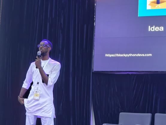 The speaker is a Black man dressed in all white in front of a slide with the link https://blackpythondevs.com