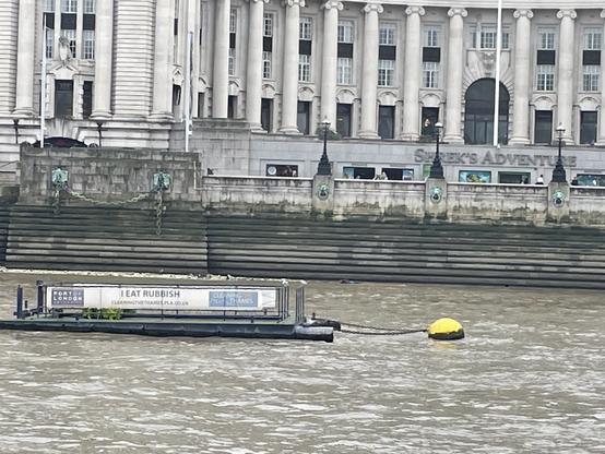 A view of the Thames, with a floating trash compactor that has a sign declaring “I EAT RUBBISH” in front of the jarringly beautiful, historic Shrek’s Adventure building.