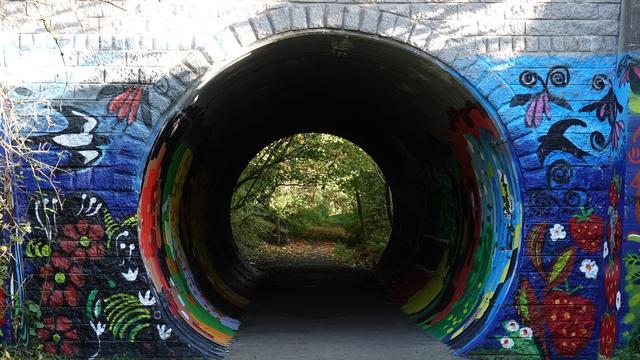An old tunnel, decorated outside with bright paintings of birds, plants and flowers. Through the tunnel a circular ringed green wooded path can be seen.