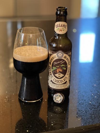 A stout glass of dark beer next to a brown bottle with a cream colored label, the center of which shows a light brown coco bean with green leaves
