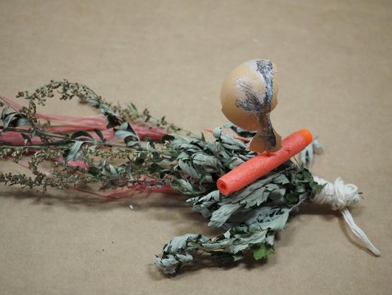 on a tan background, part of an eggshell with a clipping of a transfer drawing on it is balanced on top of a neon pink nerf bu||3t on top of dried stems gathered with a string and worn pink plastic strips underneath