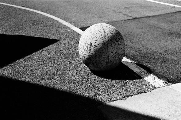Black and white photograph of a spherical concrete bollard on the edge of a bitumen paved car park. A white painted line crosses the frame from top left to bottom right.