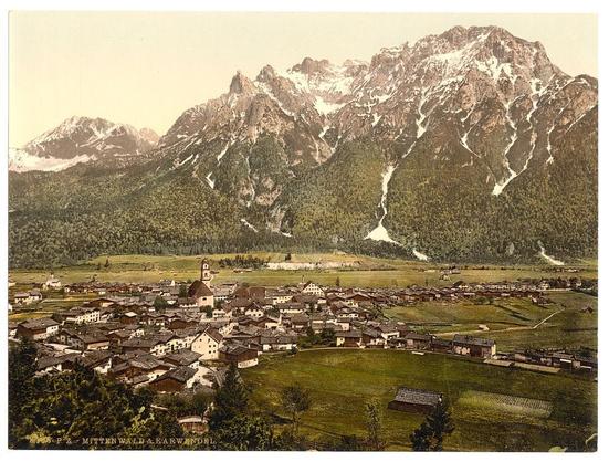 The image depicts a picturesque village nestled in the Bavarian Alps, likely dating between 1890 and 1900. The scene showcases a quaint town with traditional European architecture, consisting of wood-framed buildings topped with steeply pitched roofs made from dark shingles or tiles. Several structures feature prominent chimneys indicative of hearth-fueled heating common in the era.

The village is surrounded by lush green fields and forested hills leading up to magnificent mountains capped with snow. The mountain range serves as a majestic backdrop, highlighting the stark contrast between human habitation at lower elevations and nature's grandeur above. A church or chapel stands out within the settlement due to its tall steeple, serving both spiritual and community gathering purposes.

The image captures the essence of rural life during this period in an alpine region of Germany, providing a glimpse into historical architecture, landscape usage, and the harmonious coexistence between human settlements and nature.