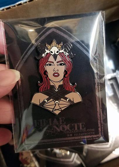 A shot of my second pin design. It's a forward facing bust of a fantasy pinup, with a crown of bird bones including a skull, and gold crystals. She has curved horns, red hair and lips, and a tan skintone. She's wearing black coiled horn earrings, a collar and harness with crescent moon accents. The pin is attached to a card bearing a silver foil tombstone and the company logo "Filiae Nocte" and is bagged in a plastic sleeve.