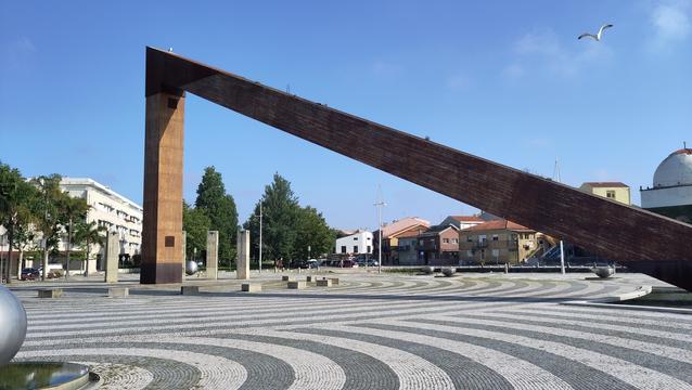 Eine große, rostige Skulptur bildet ein Dreieck. Sie ist sicher ca. 10 Meter hoch und 30 Meter breit. Es könnte ein Segel symbolisieren. Sie steht auf einem großen Platz. Das schwarz-weiße Pflaster bildet ein Wellenmuster aus konzentrischen Kreisen.