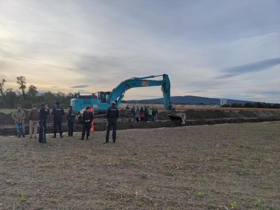 5 Polizist:innen in Uniform und weitere Personen, rechts dahinter der Bagger unter dessen Arm die Protestierenden Aktivist:innen versammelt sind, im Hintergrund eine weite Landschaft mit einem hohen Gebäude und einem Berg.