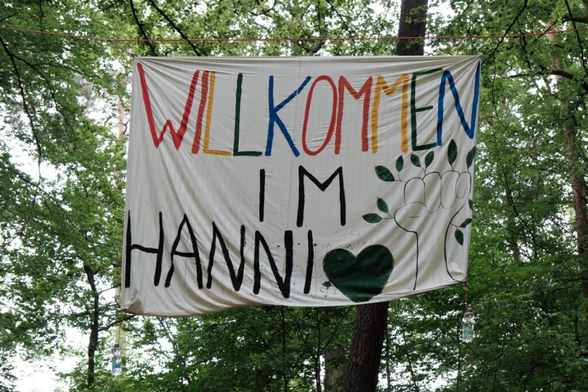 Weißer Banner aufgehängt zwischen Bäumen im Wald. Darauf steht in Bund „Willkommen“ darunter in schwarz „im Hanni“ daneben ein schwarzes Herz und daneben eine gehobene Faust (Schwarz umrandet nicht ausgemalt ) aus der Blätter wachsen