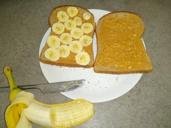 A plate with two slices of wholemeal bread, spread with peanut butter with banana slices on the left hand piece. A knife and half a banana sit in the foreground.