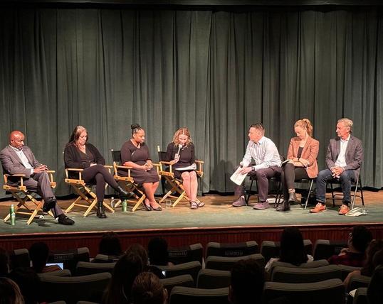Seven people, looking serious, sit on folding chairs on a stage in an auditorium.  One person with a microphone reads from a sheaf of papers.