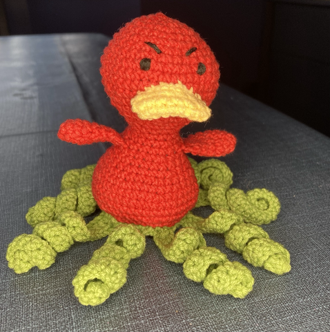 A crocheted red duck with a yellow beak and green tentacles and an evil scowl on its face!