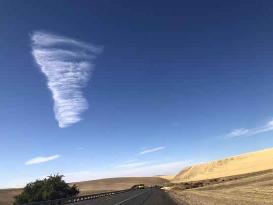 Funnel-shaped brushstroke cloud in the left upper clear blue sky with a road and a yellow truck plus 2 other cars at the foreground. Dry golden wheat stubble to the right