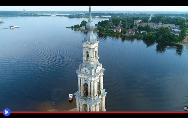 Torre ecclesiastica nello stile architettonico della Russia classica svetta stagliandosi dal centro di un bacino artificiale, ricavato dalle acque di un grande fiume. Sullo sfondo un condominio di epoca sovietica ed alcune piccole case che campeggiano sulla riva.