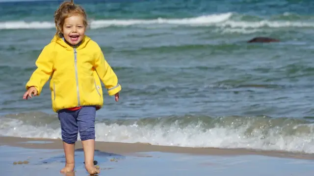 Die kleine Noemi, 6 Jahre alt, lächelt und trägt einen gelben Mantel und eine blaue Hose im Urlaub an der Ostsee