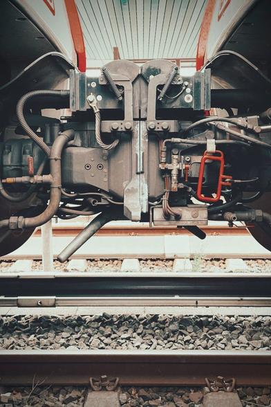 Portrait photo of the coupling between two German public transit trains. The upper half is taken up by the coupling with hitches, cables and hoses. The lower half has a hint of the tracks and gravel around them. The colors are greyish beige with a bright red handle attaches to the coupling and a bit of red from the trains‘ design.