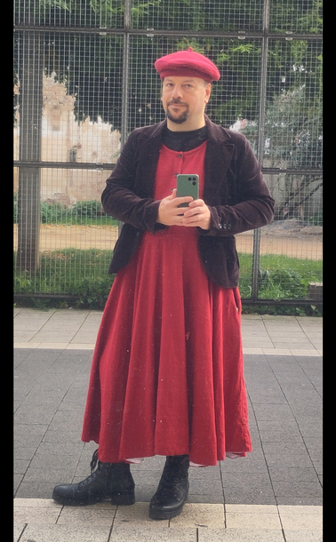 Selbstporträt in spiegelnder Scheibe. Mann mittleren Alters mit dunklem Kinnbart trägt ein langes rotes Kleid mit weitem Rockteil. Dazu eine kurze schwarze Samtjacke, eine rote Baskenmütze und schwarze Schnürstiefel.