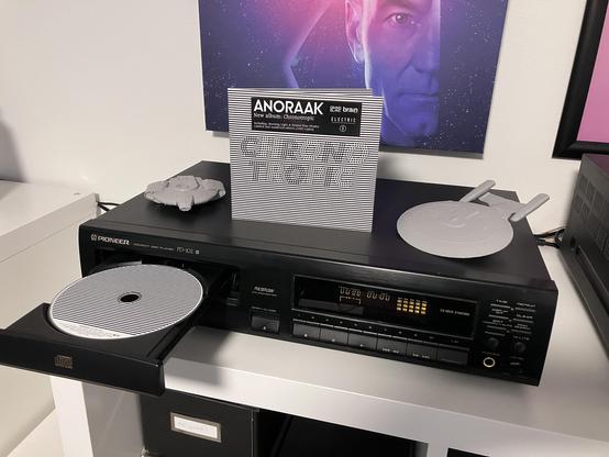 Photo of a Pioneer hi-fi compact disc player with the tray out and a CD in it. The CD digipak cover is standing on top of the player to display the album art, which is alternating black and white lines, and the album title “Chronotropic”. Also on top of the CD player are 3D printed models of the USS Defiant and USS Enterprise from the TV series Star Trek: Deep Space Nine and Star Trek: The Next Generation, respectively. On the wall can be seen the lower part of a poster from the movie Star Trek: Generations.
