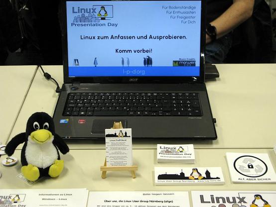 Foto: Ein Notebook mit Linux-Installation mit Hinweis auf unseren nächsten Linux Presentation Day 2024.2 am 16. November wieder in Kooperation mit dem Fablab Region Nürnberg in der Schwabacher Str. 512.  Auf dem Tisch links ein kleiner Tux, in der Mitte unsere Visitenkarten und diverse Aufkleber.