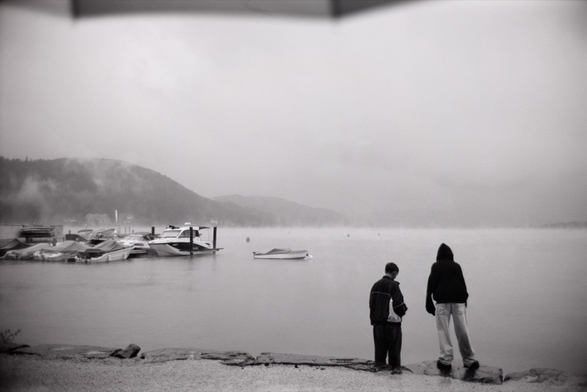 The image shows two people standing on the edge of a misty, calm lake. The scene is set in a grayscale, creating a moody and serene atmosphere. On the left, several small boats are docked at a pier, and faint mountains are visible in the background, partially obscured by low-hanging clouds and fog. The two figures, one shorter and one taller, stand with their backs to the camera, gazing out at the lake. Both are dressed in hooded jackets, suggesting it might be cold or rainy. The scene feels quiet and introspective, with an umbrella partially visible at the top, hinting at rain.