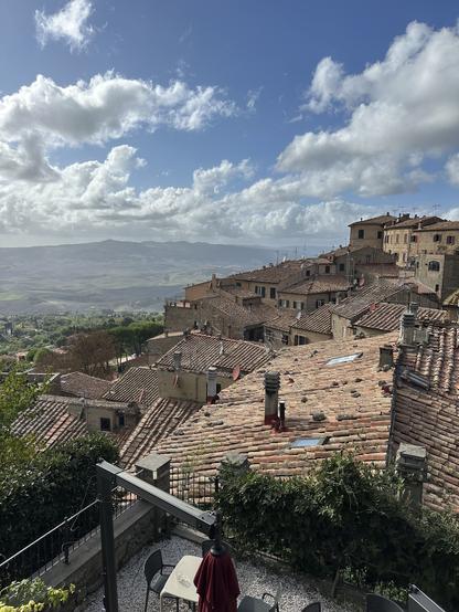 A scenic view of a hillside village with terracotta rooftops under a partly cloudy sky. The foreground features a small patio area with a table and chairs, surrounded by greenery, while rolling hills extend in the background.