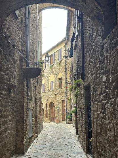 A narrow cobblestone alleyway flanked by rustic stone buildings, featuring flower pots and a lantern. The archway overhead frames a glimpse of a sunlit street with wooden doors and shuttered windows.