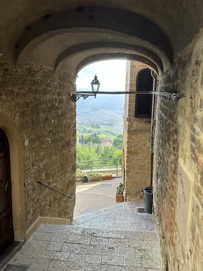 A stone archway leading to a staircase with a view of a lush landscape. A vintage lantern hangs above, with potted plants visible at the bottom of the stairs.