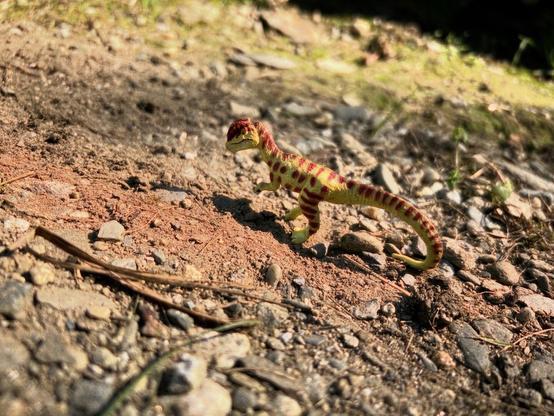 A green Dilophosaurus with brown spots standing in an arid, rocky plain as the sun beats down on them.