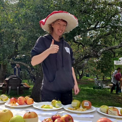 Ella, at heritage apple tasting station, giving a thumbs up & wearing a red & white mushroom hat.