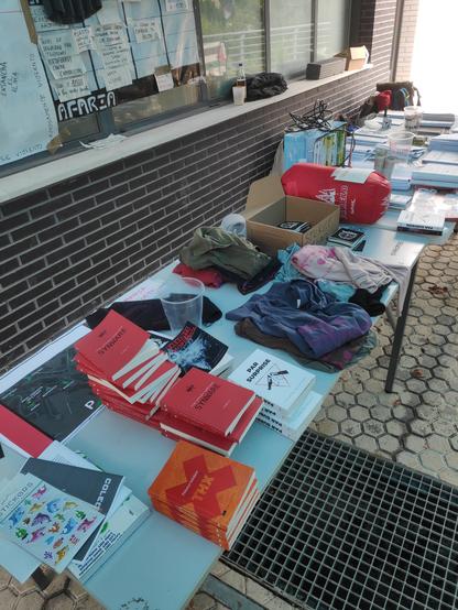 Foto del infopoint con libros, más libros, pegatinas, camisetas, un saco de dormir perdido, fanzines y mochilas al fondo