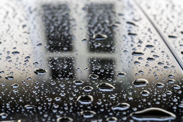 Das Bild zeigt eine Nahaufnahme eines Autodachs, das von vielen Wassertropfen bedeckt ist. Die Tropfen variieren in Größe und Form und reflektieren das Licht, was ihnen eine kristallklare, glänzende Erscheinung verleiht. Die Oberfläche scheint ein Fahrzeug oder eine Glasfläche zu sein, und im Hintergrund sind unscharfe vertikale Formen eines Fensters zu erkennen, die möglicherweise Gebäude oder eine reflektierte Struktur darstellen. Der Fokus liegt auf den scharfen Details der Wassertropfen im Vordergrund, während der Hintergrund weich und unscharf bleibt, was dem Bild eine Tiefe und Textur verleiht.